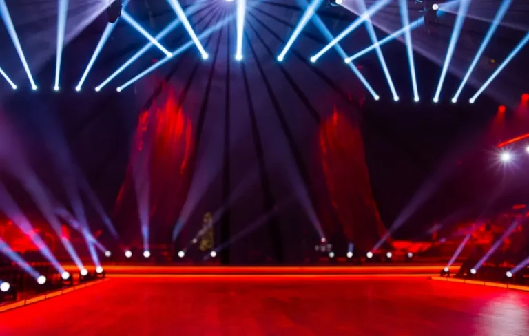 A big stage viewed from performers’ perspective, w/ red lighting except for bright white lights along the stage edge